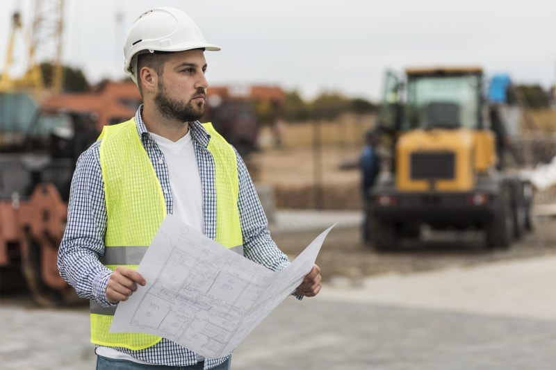 Man in a yellow construction vest and white hard hat holding road plans with heavy duty road construction equipment in the background.