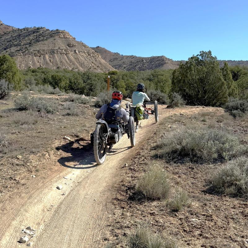 Two people are biking on the trails at North Fruita Desert. They are using specially modified electric motor assisted tricycles called adaptive mountain bikes.
