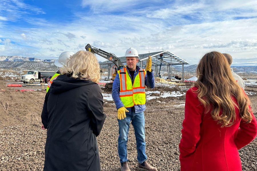 Man stands in front of construction wearing bright green and orange construction vest and two women face the construction site. 
