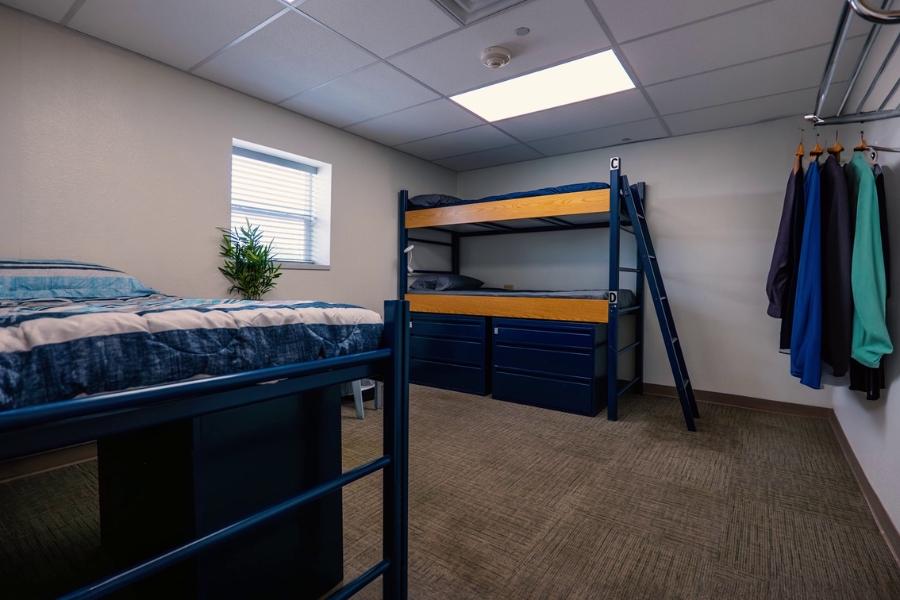 Dormitory with a window, a blue bunk bed, a blue single bed, and a nightstand with a plant on it.