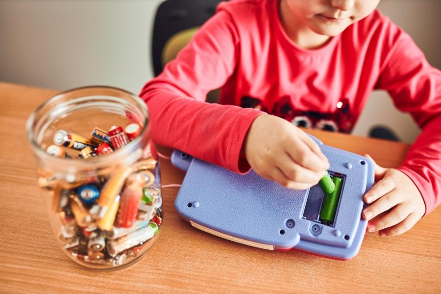 A child putting green batteries in purple toy next to a glass jar of many variations of batteries. 