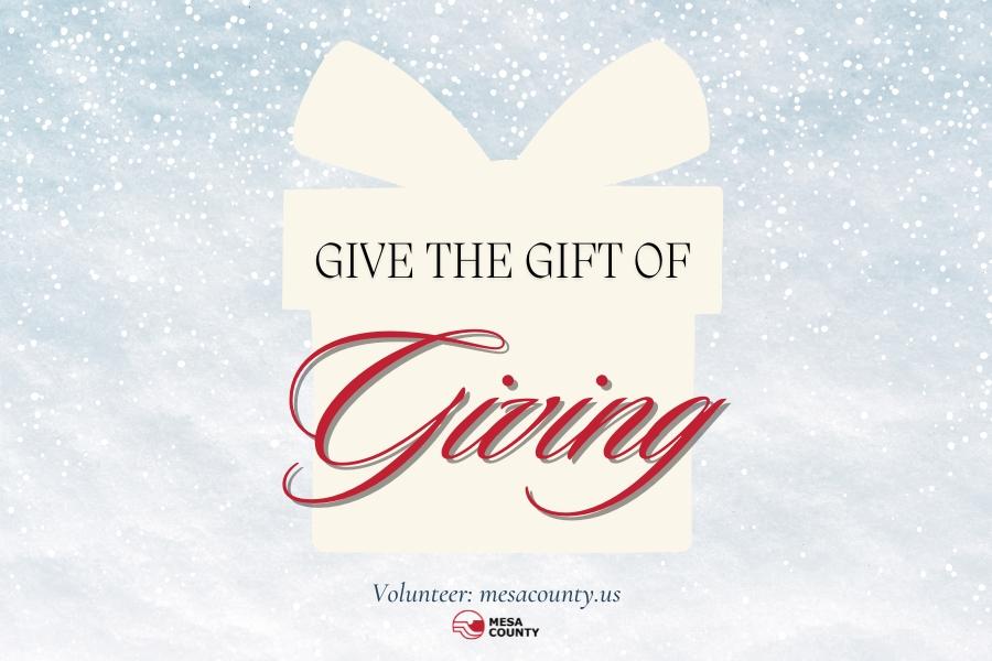 Light blue snowy background with white present element and black and red text reading, "Give the Gift of Giving Volunteer: mesacounty.us," and the red and black Mesa County logo. 