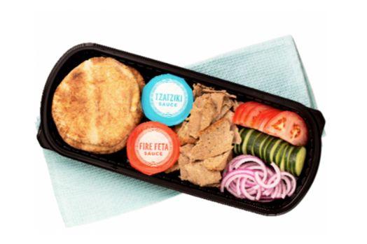 Black, plastic container containing pita bread, slices of meat, tomato slices, cucumber slices, red onions, tzatziki sauce, and fire feta sauce.