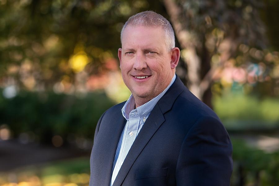 Man (Todd Hollenbeck) in blue suit jacket smiling for headshot in front of blurred trees.