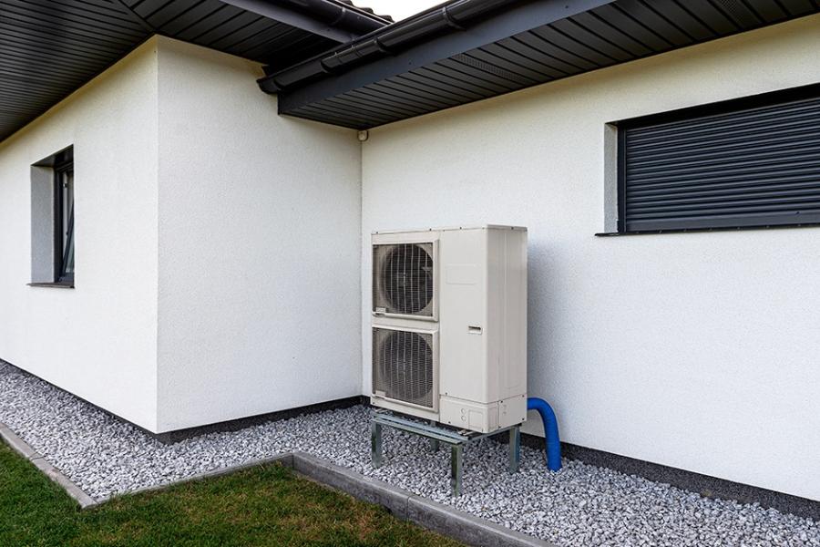 A heat pump sits outside of a single-story house, placed on a platform elevated about a foot off of the ground.