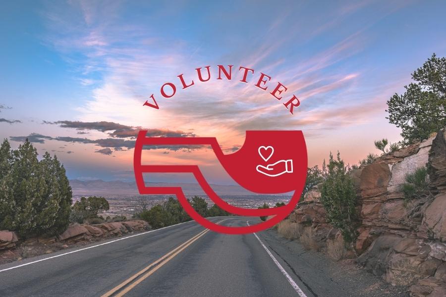 Empty road with two yellow lines in the middle in between rocks and trees on the Colorado National Monument with red Mesa County logo, white hand below heart element, and red text reading volunteer. 