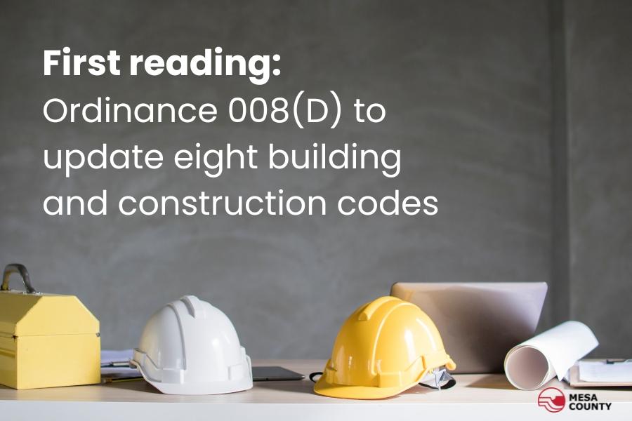 A yellow tool box, white and yellow construction hats, rolled up paper, and silver computer sit on a white desk with white text reading, First reading: Ordinance 008(D) to update eight construction codes with the red and black Mesa County logo at the bottom. 