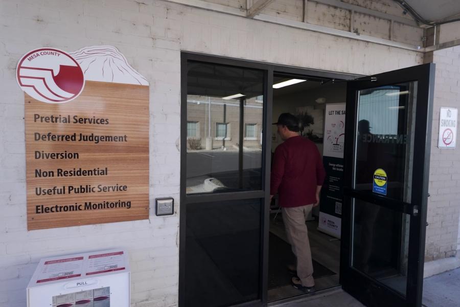 Man walking into building with sign including Mesa County logo and black text reading, Pretrial Services Deferred Judgement Diversion Non Residential Useful Public Service Electronic Monitoring. 