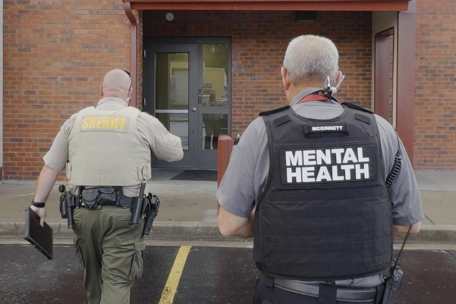 A sheriff’s deputy and a mental health professional walk side by side toward a building entrance. The deputy wears a tan vest labeled "SHERIFF," and the clinician wears a black vest labeled "MENTAL HEALTH." 
