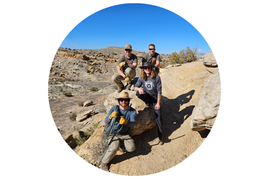Four members of the Mesa County Trail Crew pose together on a desert trail surrounded by rocky terrain and blue sky.