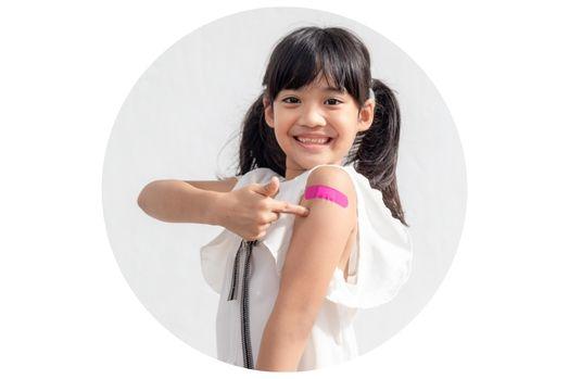 A young person wearing a white shirt smiles and points to a pink bandage on her arm.