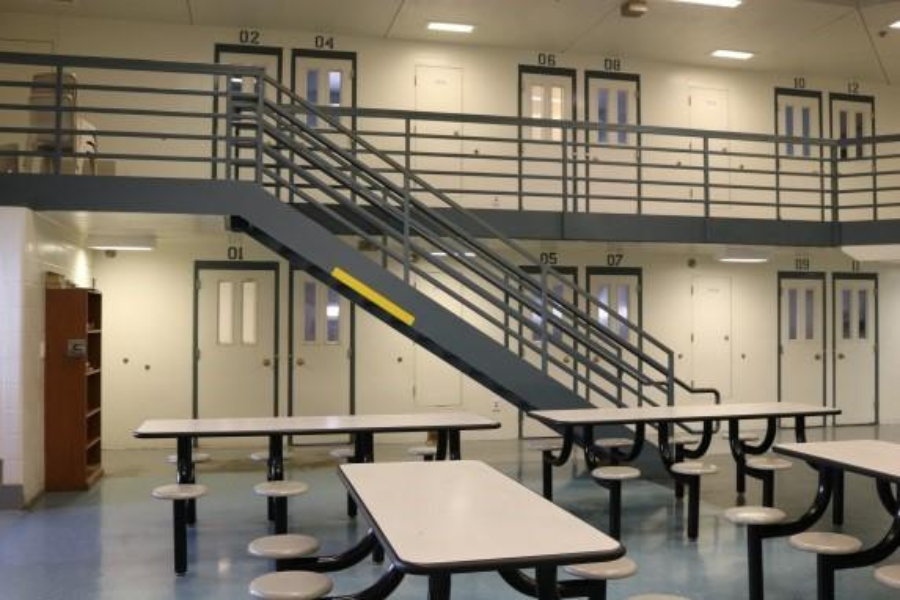 Interior of a detention facility housing unit with tables, benches, stairways and numbered cell doors on two levels.